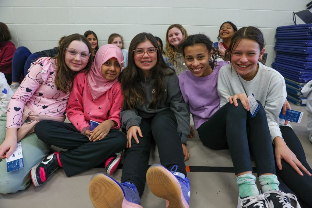 Group of young girls sitting on floor in school gymnasium, posing for picture.