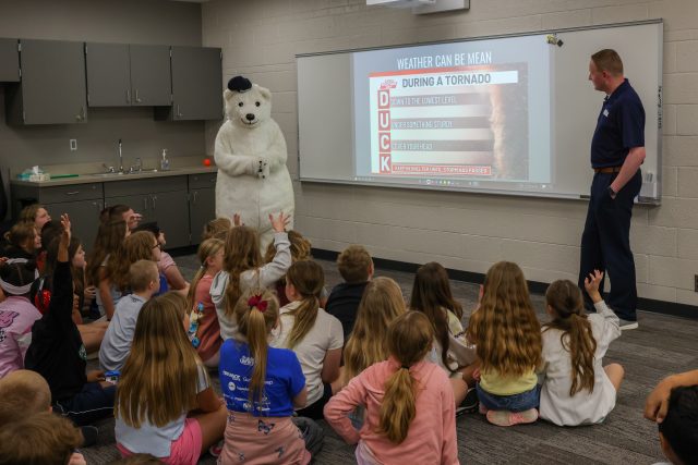 An animated bear character stands in front of a classroom full of children, who are watching and interacting with it.