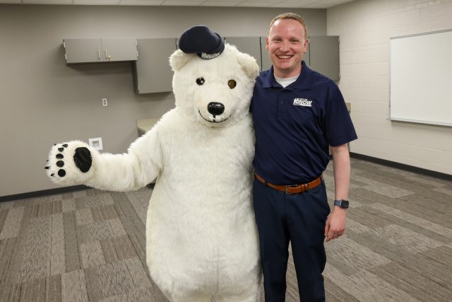 Man standing next to a large white teddy bear mascot in an indoor setting.