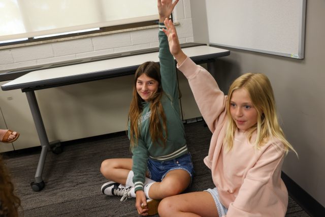 Two young girls in a classroom setting, one raising her hand while the other is sitting on the floor, both appear to be