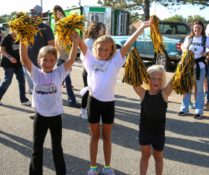 Three young girls holding pom poms, standing on a street next to a car and a truck.