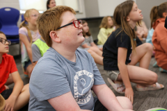 Boy in grey shirt sitting on floor in front of group of people.