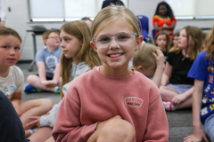 Girl wearing glasses smiling at camera in classroom setting.