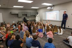 In a classroom setting, an adult man stands in front of a group of children, gesturing towards a presentation on the screen