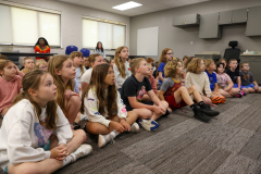 Children sitting in a classroom setting, facing towards the front of the room where an adult is speaking.